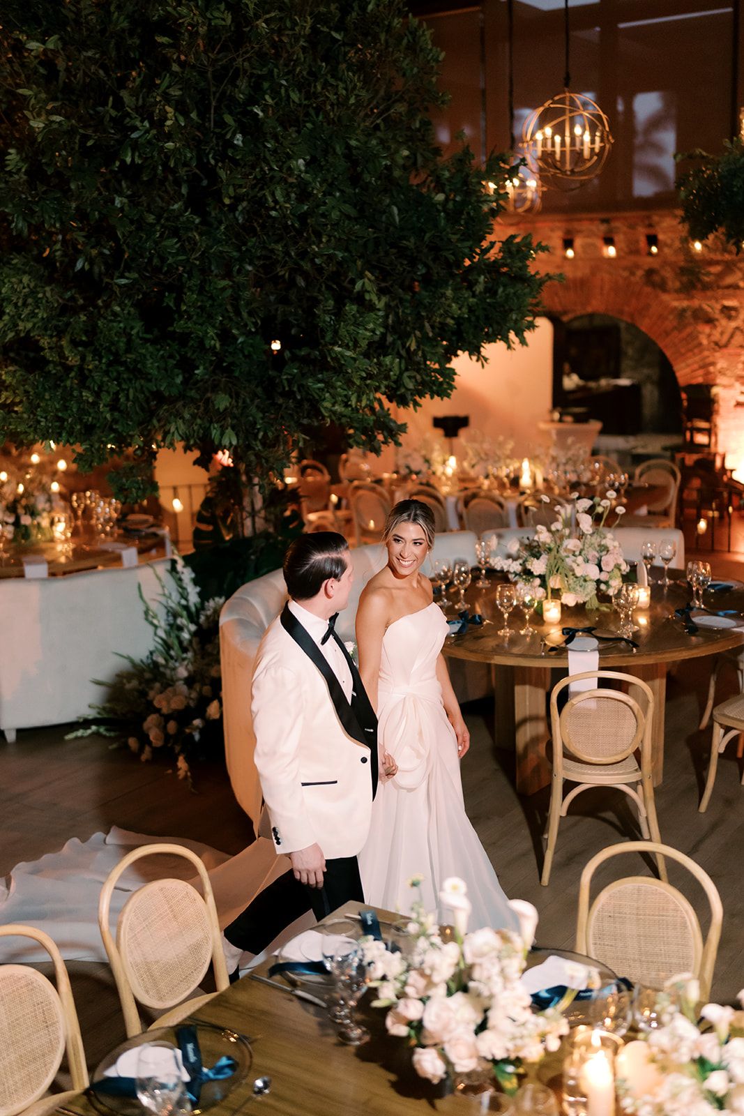 Bride and groom share a romantic moment at elegant wedding reception with candlelight, floral arrangements, and lush greenery.