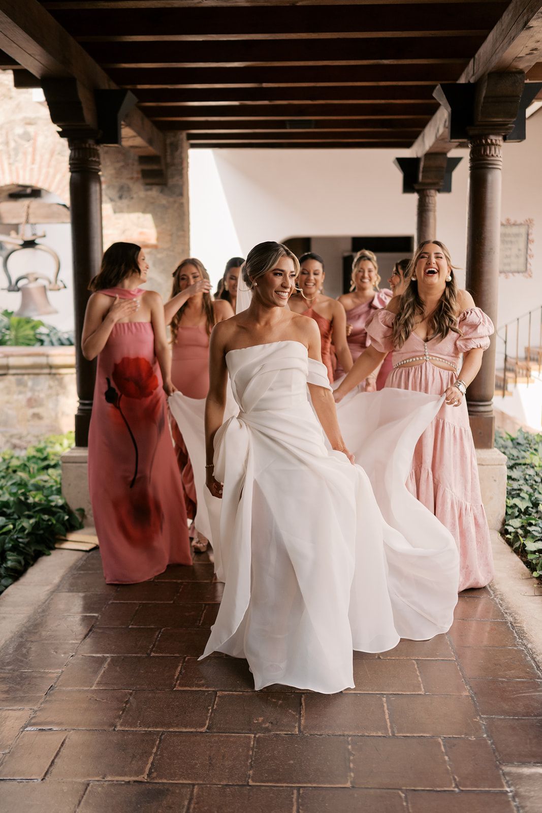 Bride in white gown walks with laughing bridesmaids in pink dresses along a Spanish-style covered walkway.