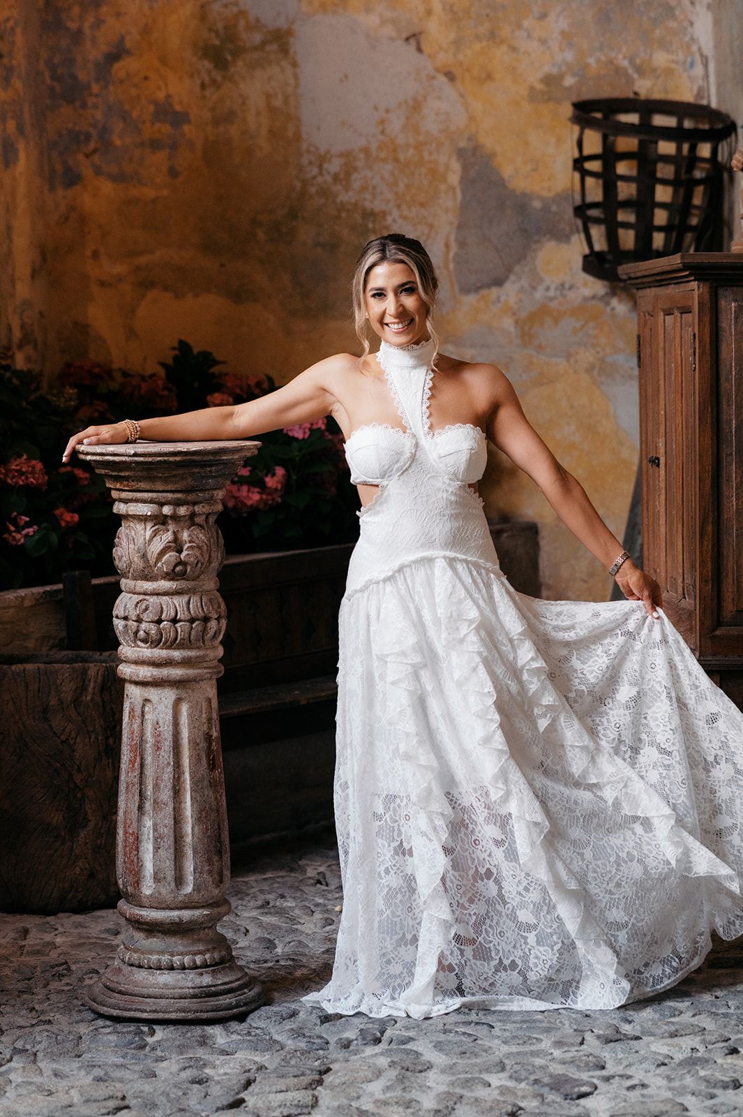 Smiling bride in white lace halter wedding gown poses beside stone pedestal in rustic venue.