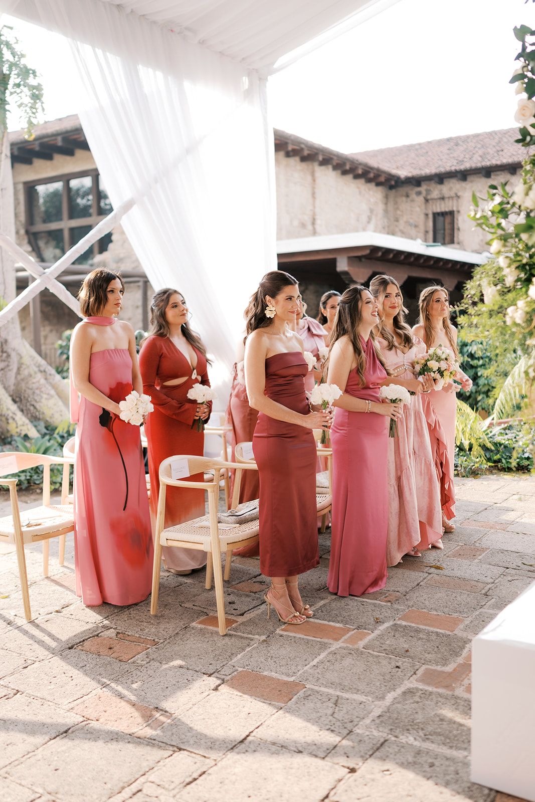 Bridesmaids in mismatched pink and red gowns holding bouquets stand at an outdoor wedding ceremony.