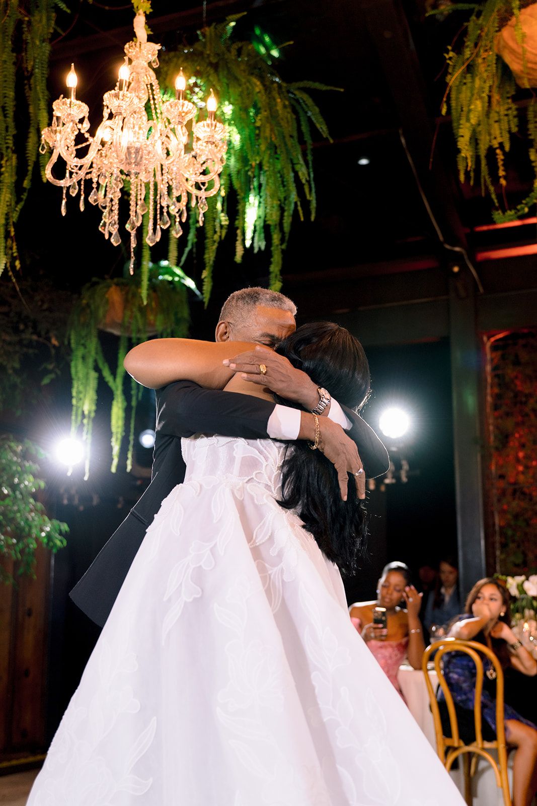 Bride and groom share first dance under chandelier and lush greenery at elegant wedding reception.