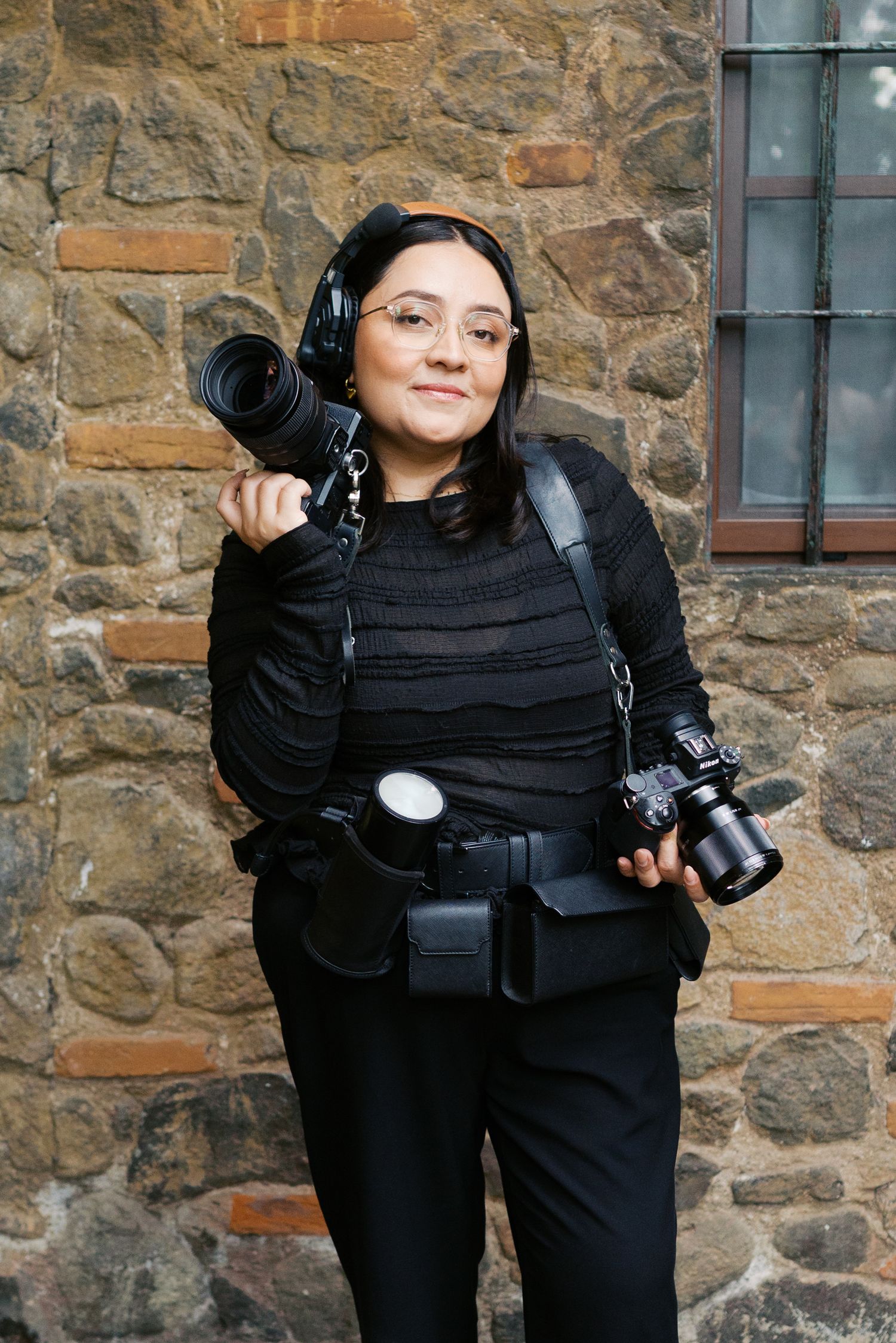 Female photographer in black outfit holding DSLR cameras against rustic stone wall backdrop.