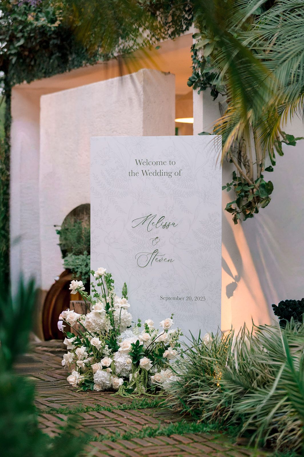 Elegant outdoor wedding welcome sign surrounded by lush greenery and white floral arrangements for Melissa and Steven.