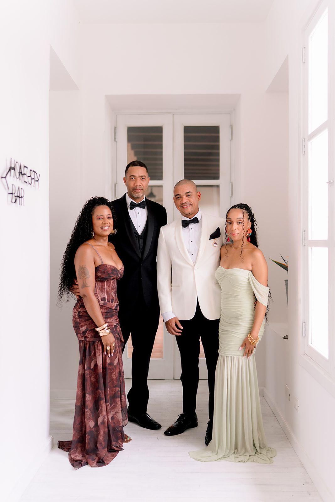 Two grooms in tuxedos pose with two women in elegant gowns in a bright white hallway at a wedding.