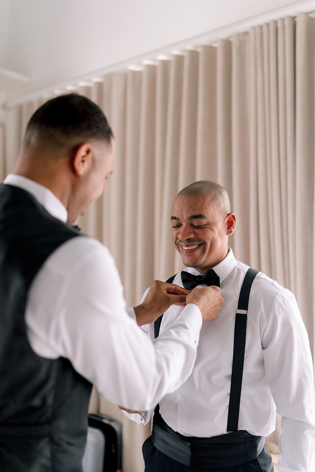 A smiling groom in suspenders and bow tie gets help from his groomsman while preparing for his wedding day.