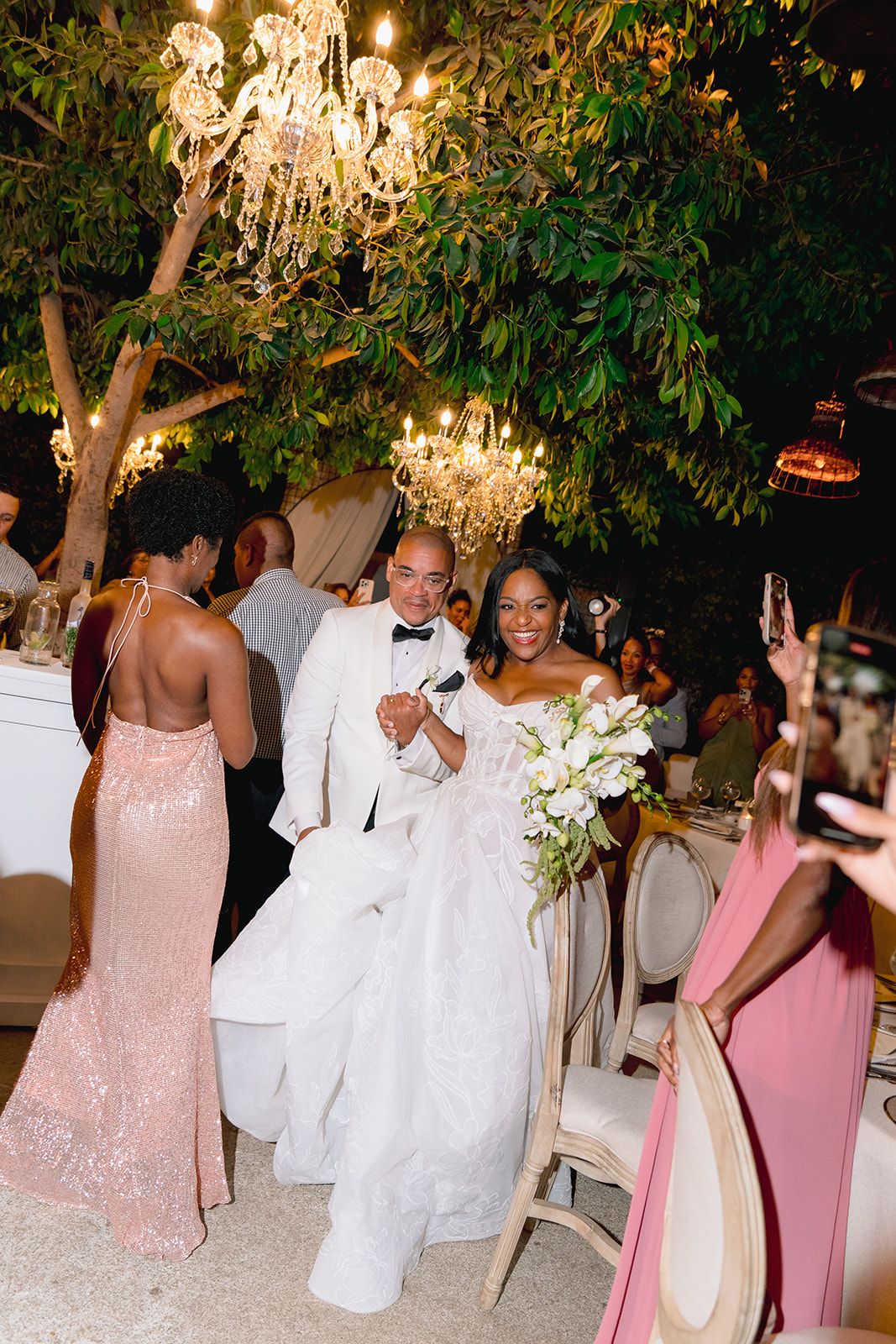 Bride in white gown and groom in white tuxedo celebrate at outdoor wedding reception under glowing chandeliers.