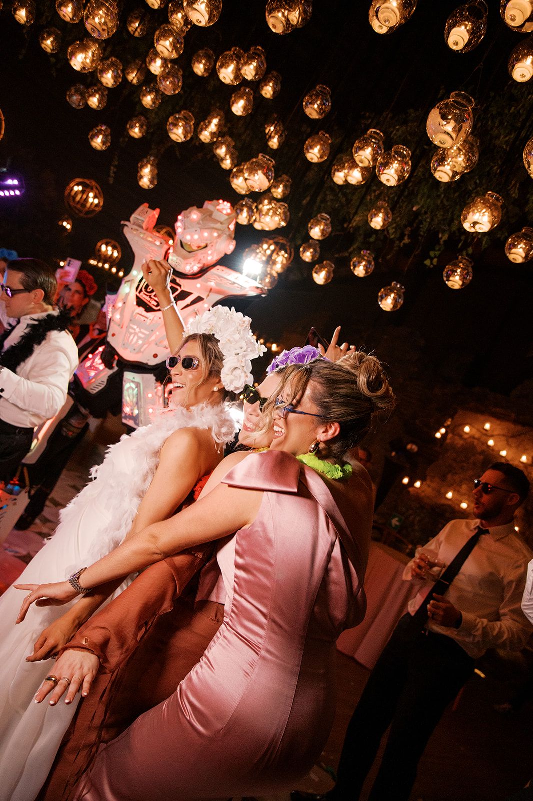 Two women in pink and white dresses dance under warm Edison bulb lights at an elegant outdoor wedding reception.