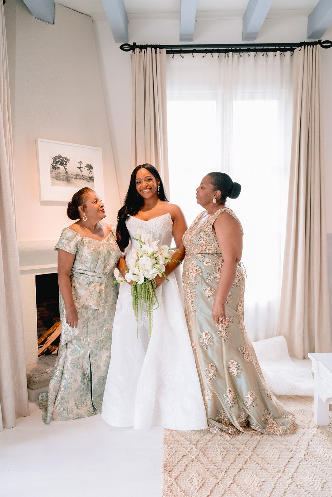 Bride in white gown holds bouquet flanked by two women in floral dresses, smiling in elegant bright room.