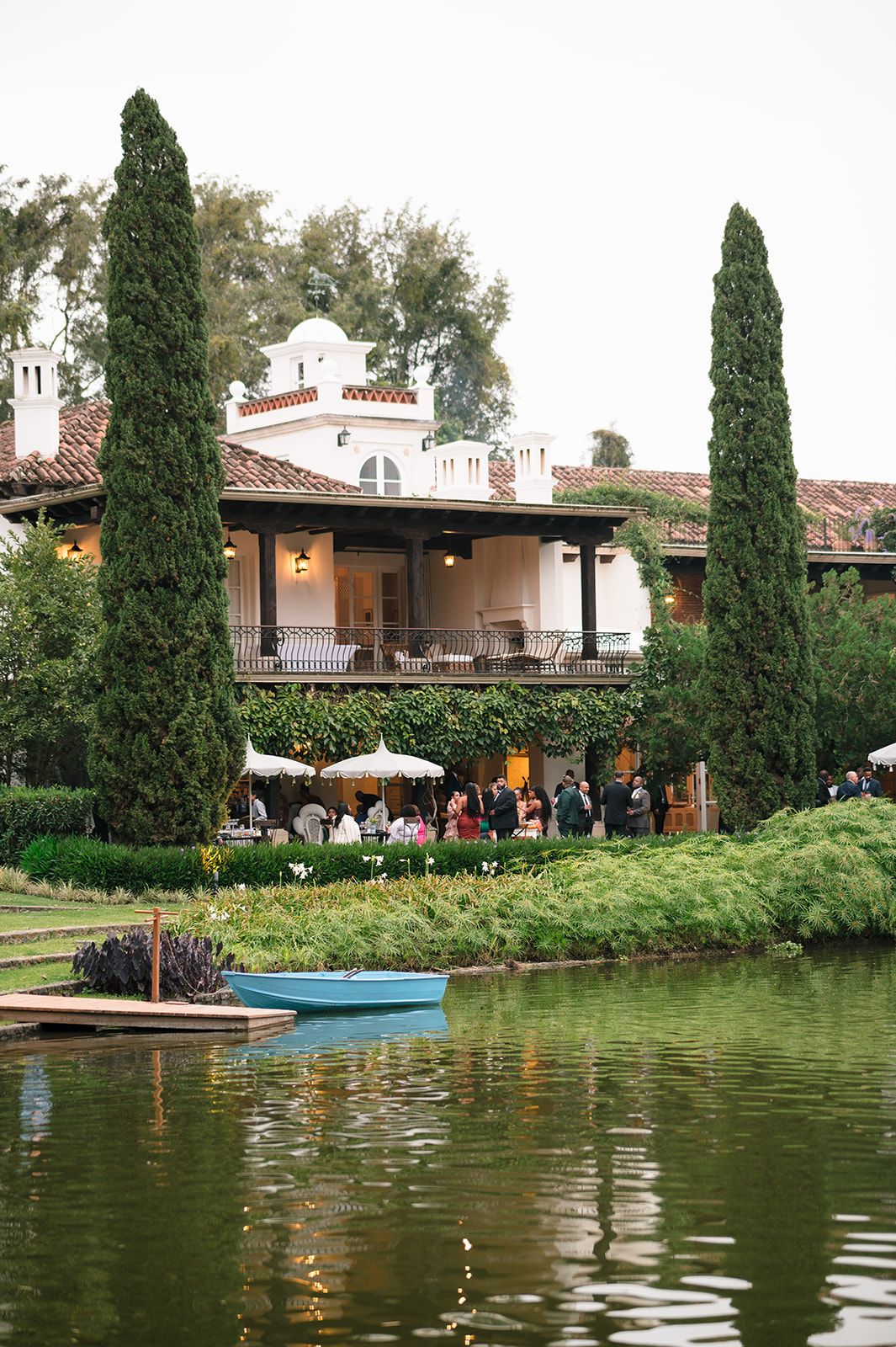 Spanish-style villa with cypress trees reflected in a serene pond, with guests gathered on the patio during an outdoor event.