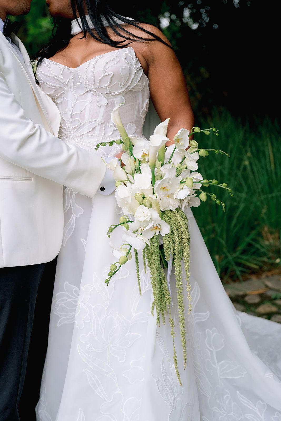 Bride in white strapless gown holds elegant cascading bouquet of white orchids and greenery while embracing groom in white tuxedo.