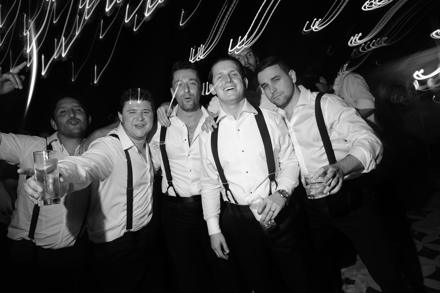 Black and white photo of five men in white shirts and suspenders celebrating at a wedding reception party.