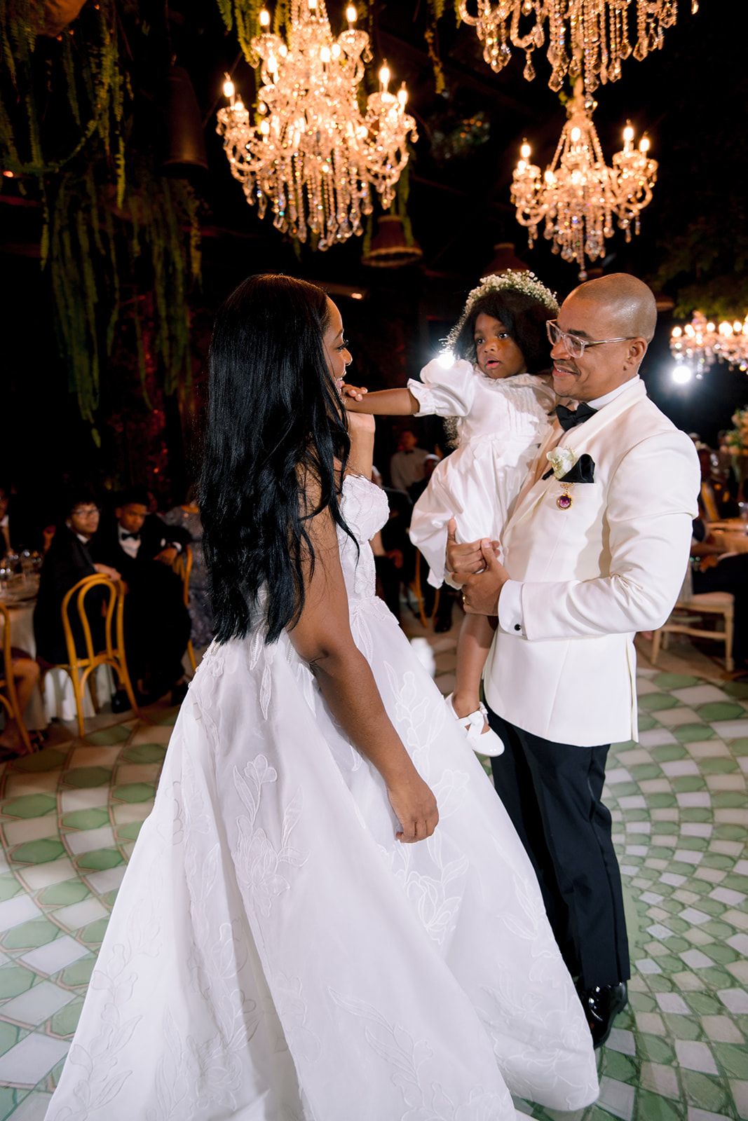 Bride and groom in white attire share first dance while holding their young child under glowing chandeliers at wedding reception.