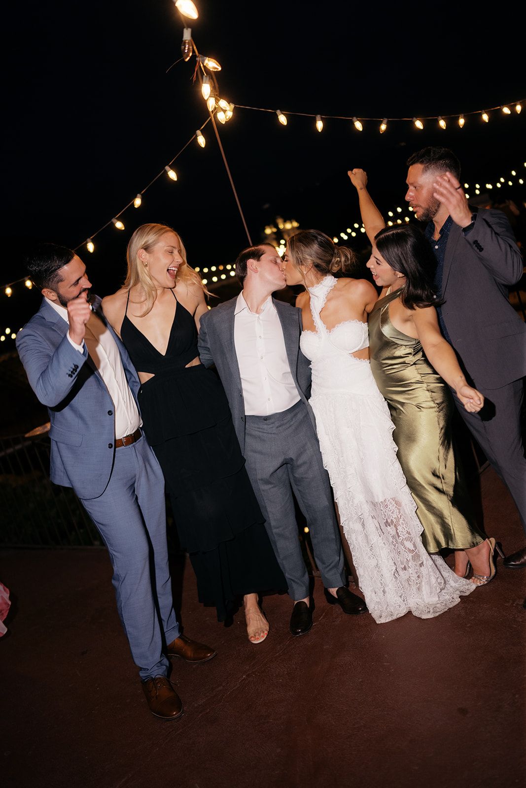 Wedding couple kissing surrounded by celebrating guests under string lights at an outdoor evening reception.