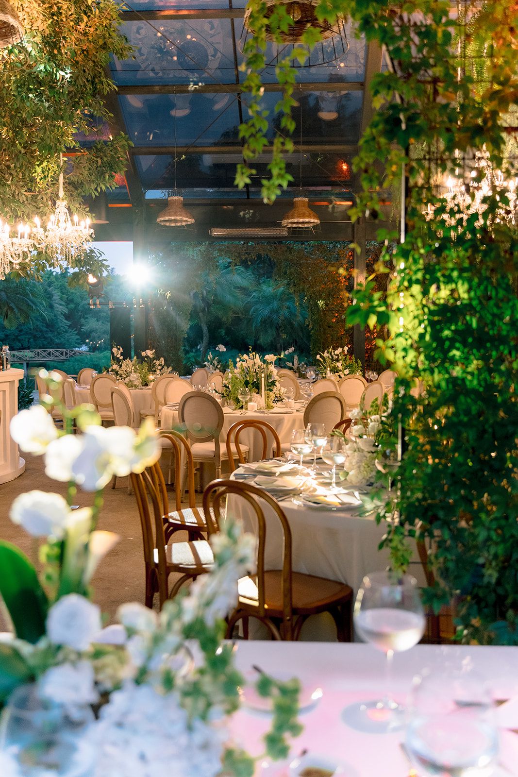 Elegant indoor wedding reception with lush greenery, white floral centerpieces, and candlelit tables in a greenhouse venue.