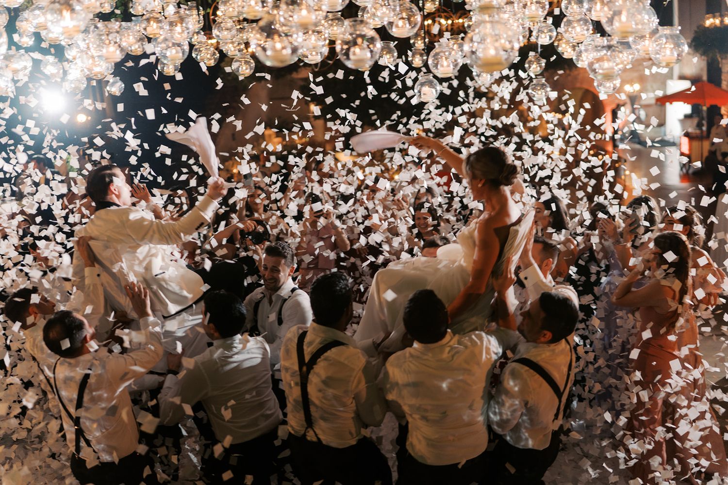 Bride and groom lifted by guests amid swirling white confetti at a festive wedding reception celebration.