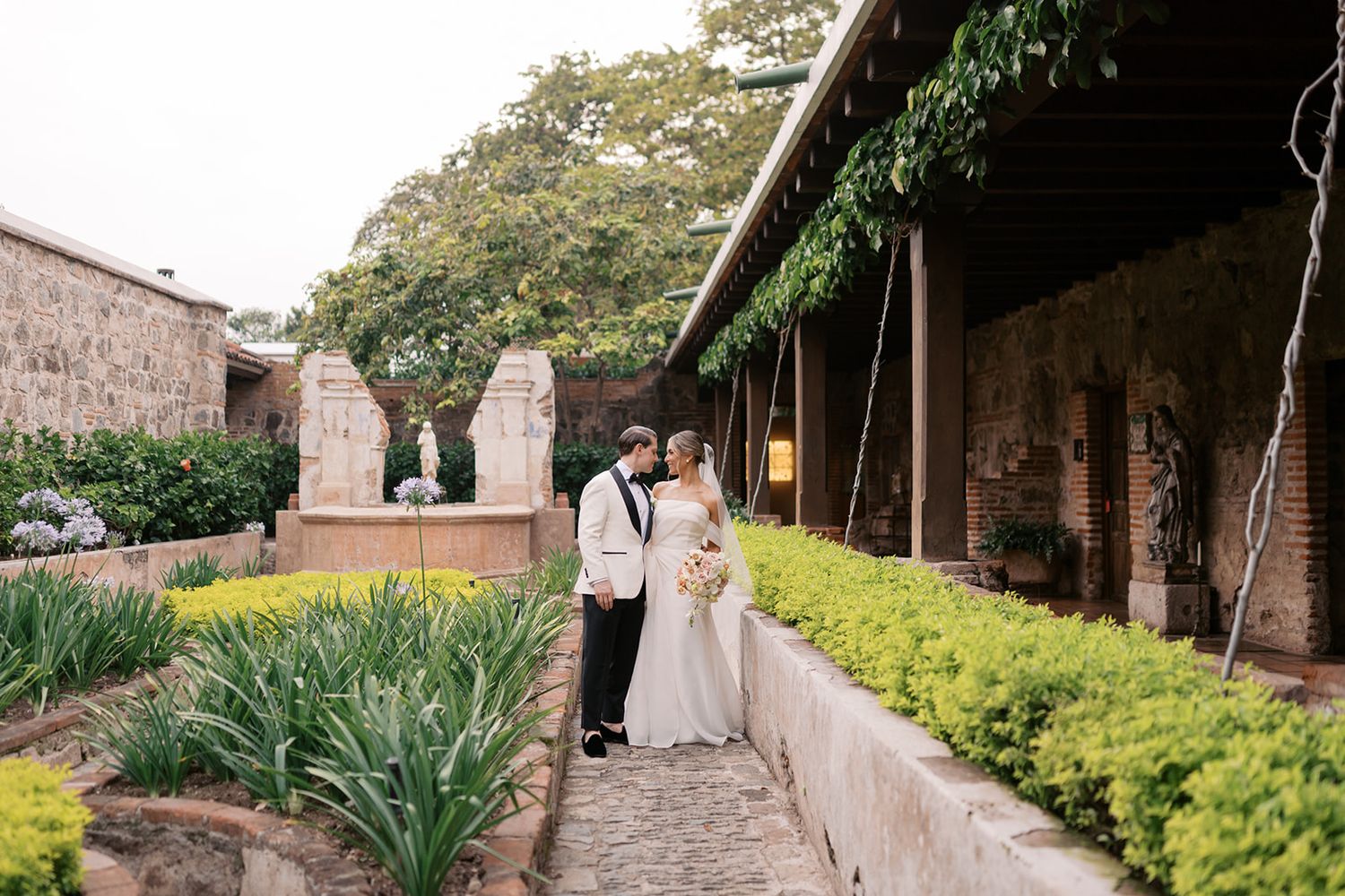 Bride and groom pose in a lush garden courtyard with stone architecture and greenery at an elegant outdoor wedding venue.
