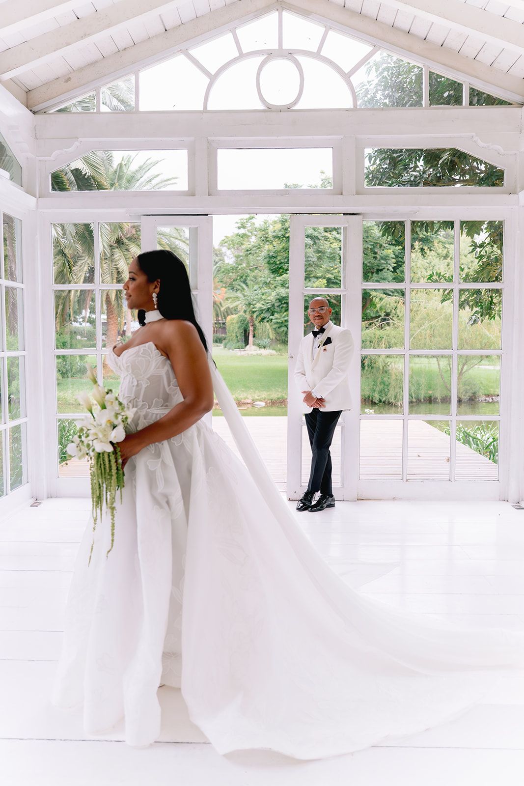 Elegant bride in white gown holding calla lily bouquet, groom in white tuxedo standing in bright glass conservatory.