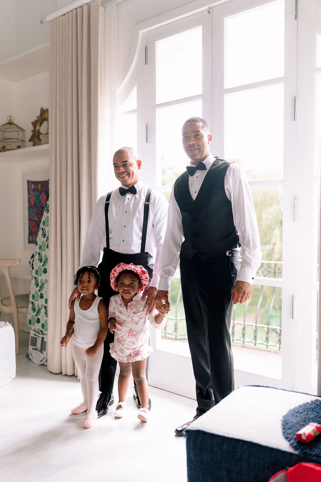 Two grooms in formal attire with bow ties pose with two young girls in a bright white room on their wedding day.