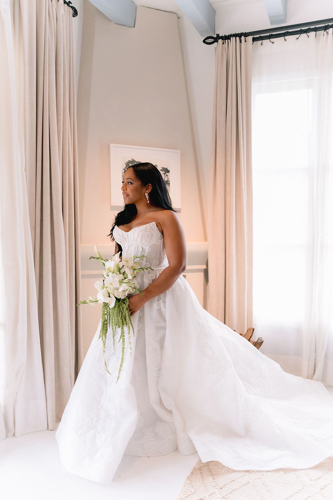 Elegant bride in strapless white gown holding lush white floral bouquet, standing in bright room with draped curtains.