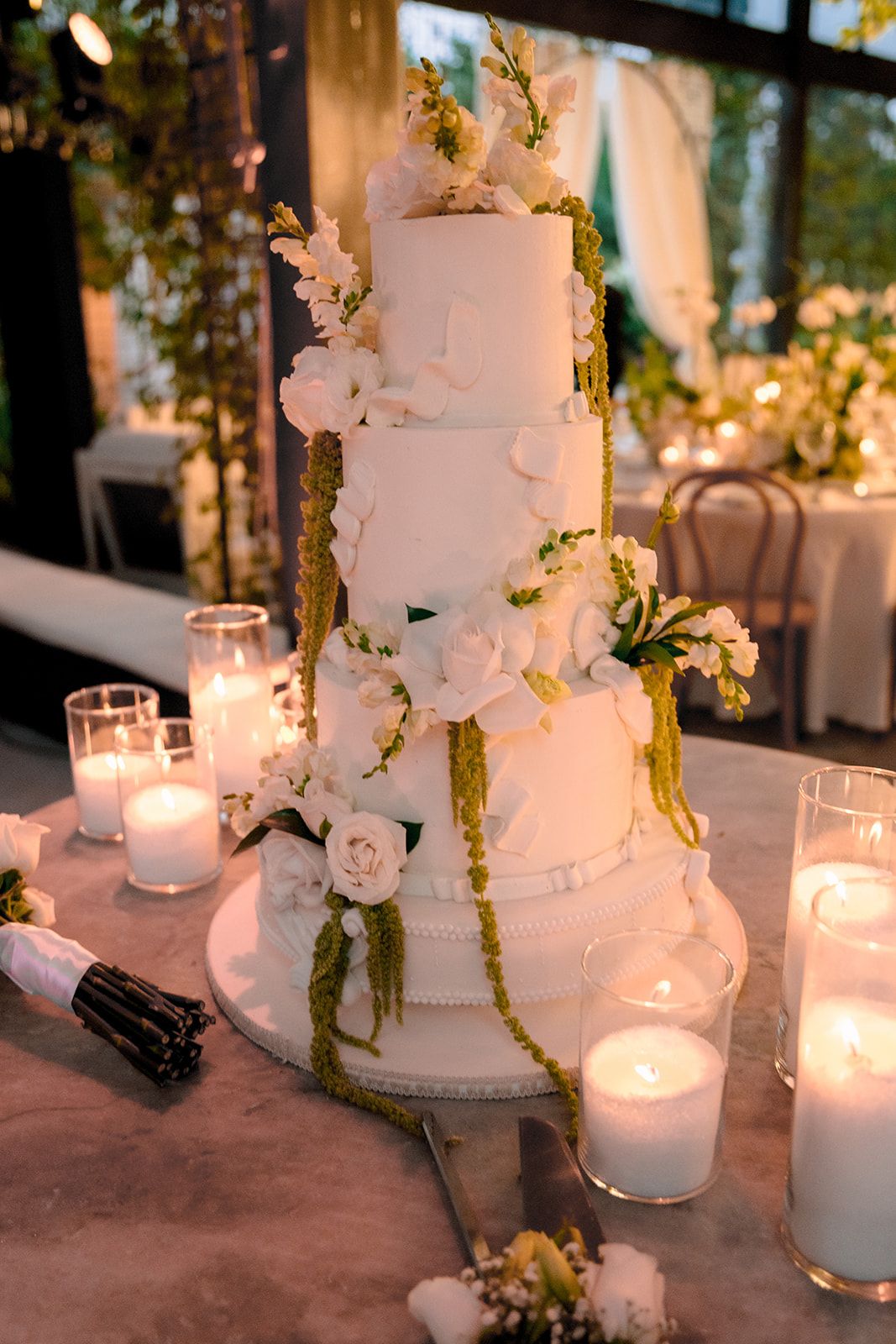 Elegant four-tier white wedding cake adorned with white flowers and green garland, surrounded by glowing candles on a marble table.