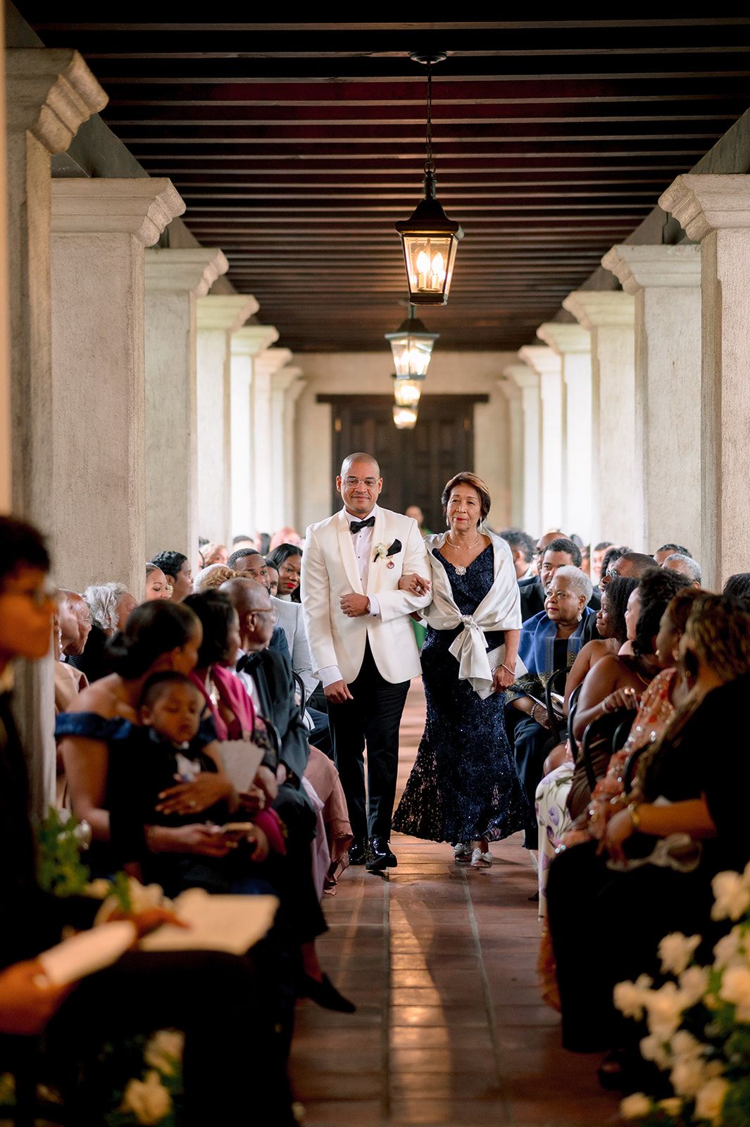 Groom in white tuxedo escorts a woman in navy gown down wedding ceremony aisle in elegant columned venue.
