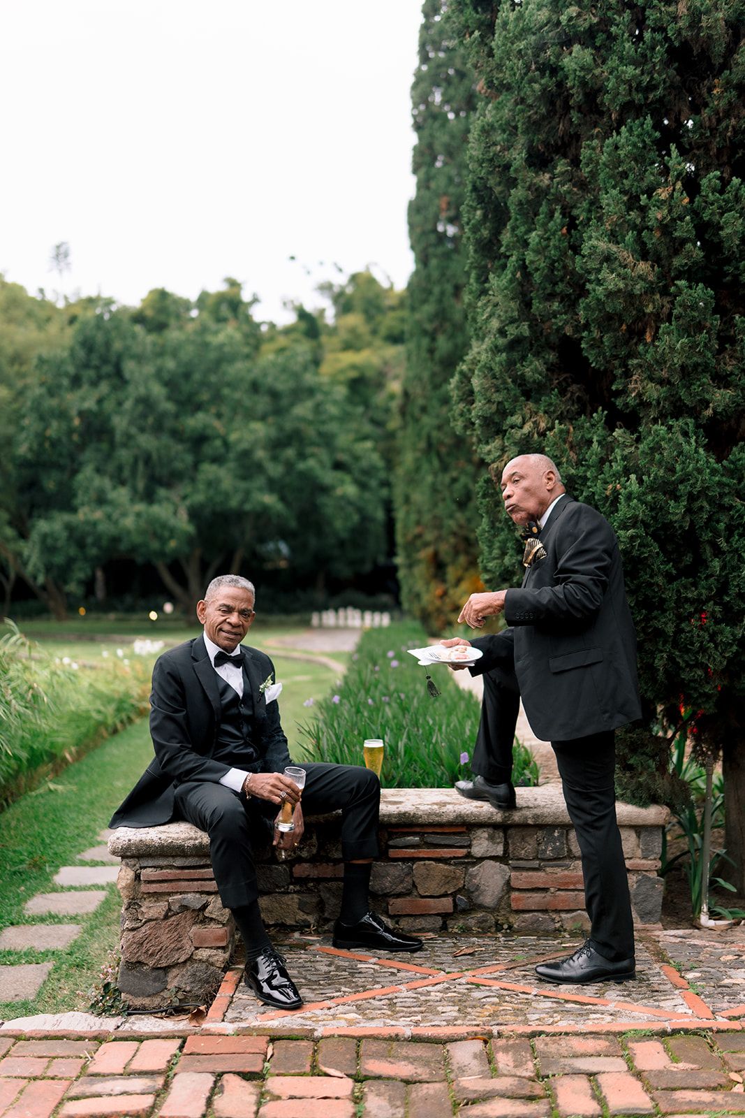 Two men in tuxedos converse outdoors near stone benches and tall cypress trees at a formal garden event.