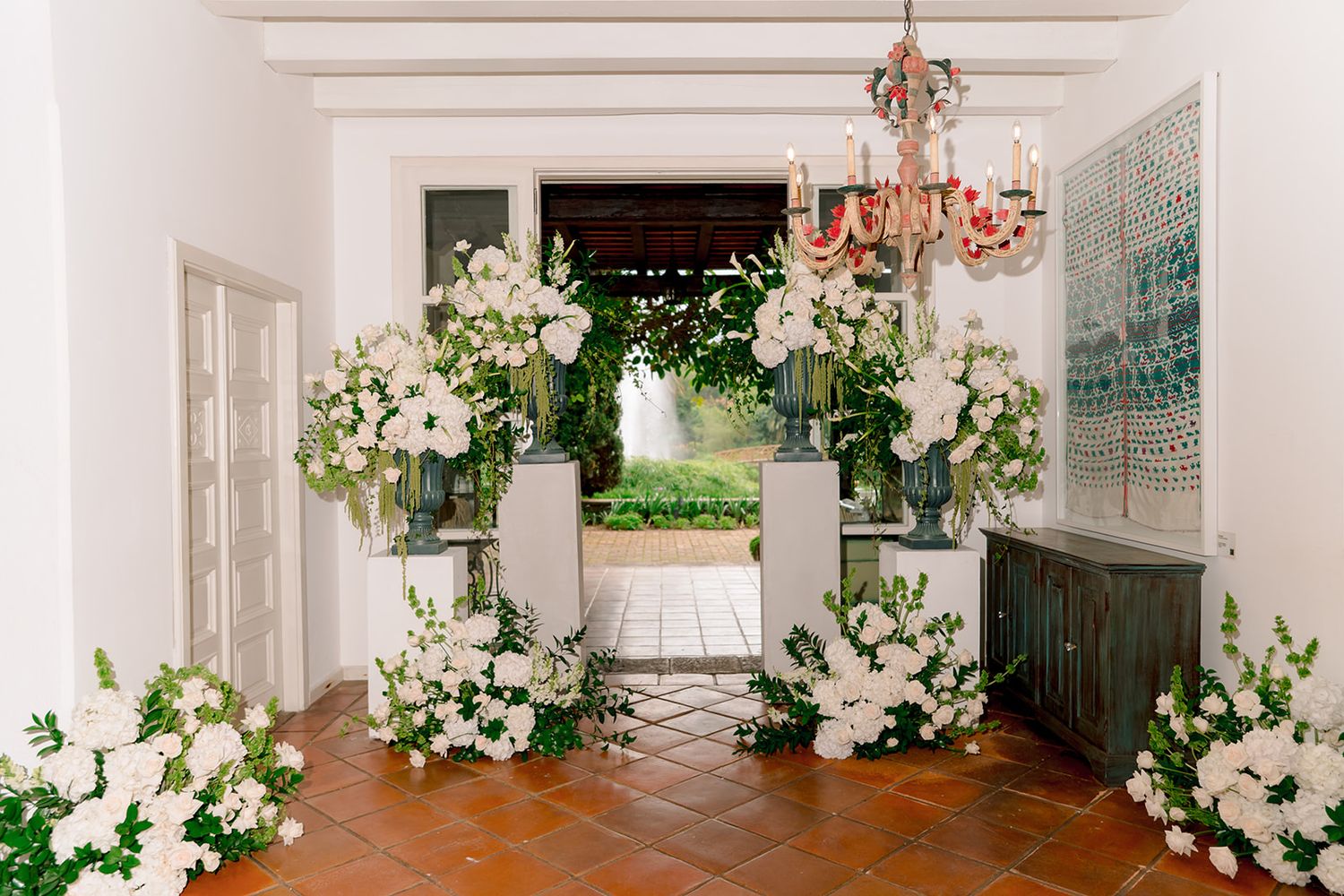 Elegant wedding venue entrance adorned with lush white floral arrangements on pedestals, chandelier, and terracotta tile floors.
