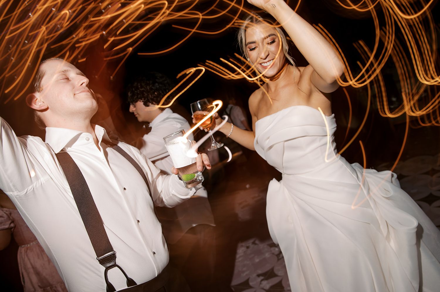 Bride and groom dancing joyfully at wedding reception with swirling light trails in background.