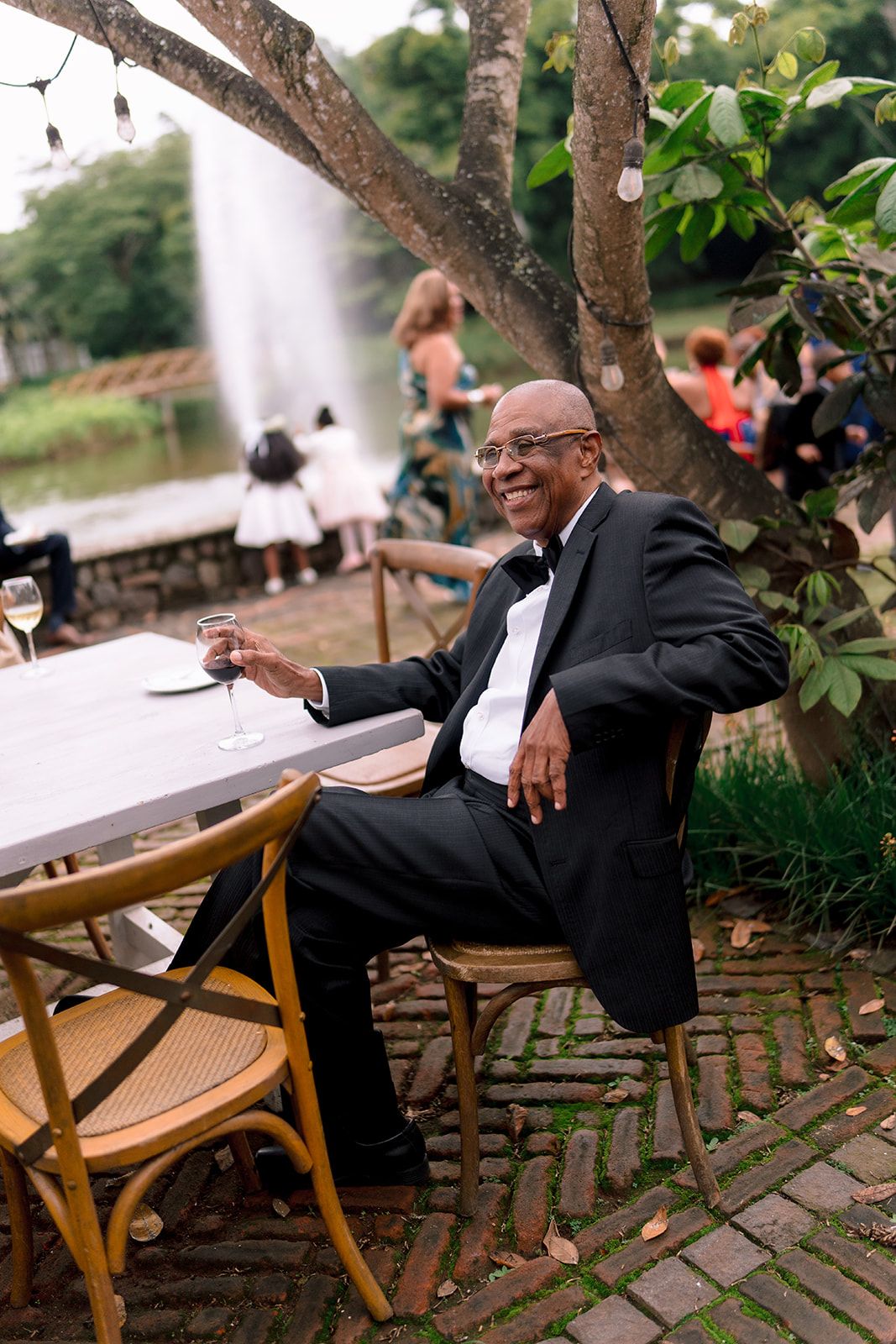 Smiling man in tuxedo sitting at outdoor wedding reception table holding champagne glass near fountain.