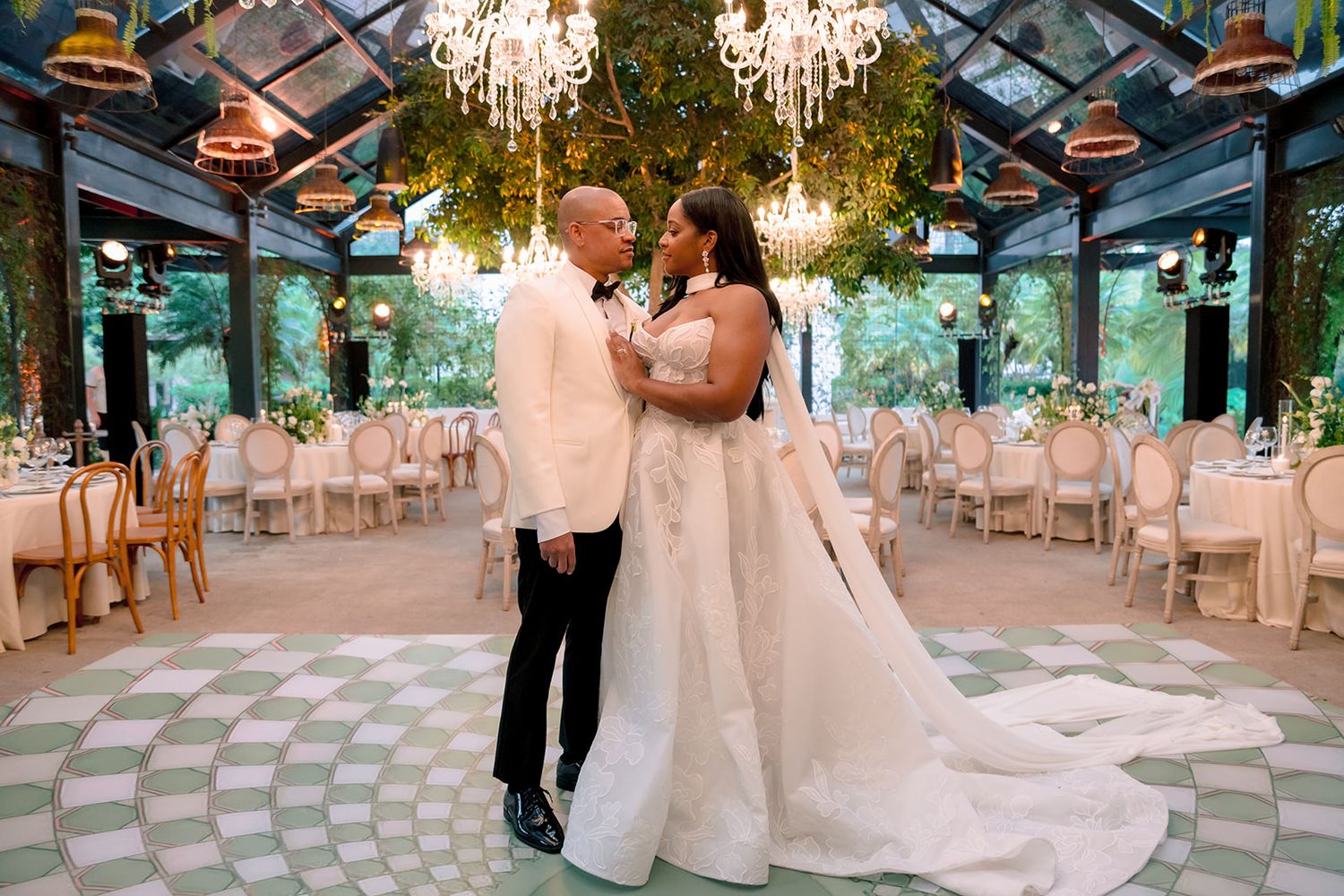 Elegant wedding couple embracing in a stunning greenhouse venue with chandeliers, lush greenery, and white reception tables.