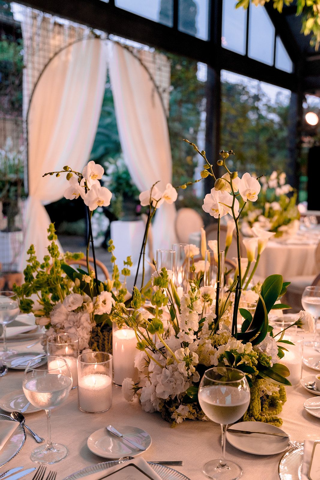 Elegant wedding reception table with white orchids, candles, and greenery centerpiece in a greenhouse venue.