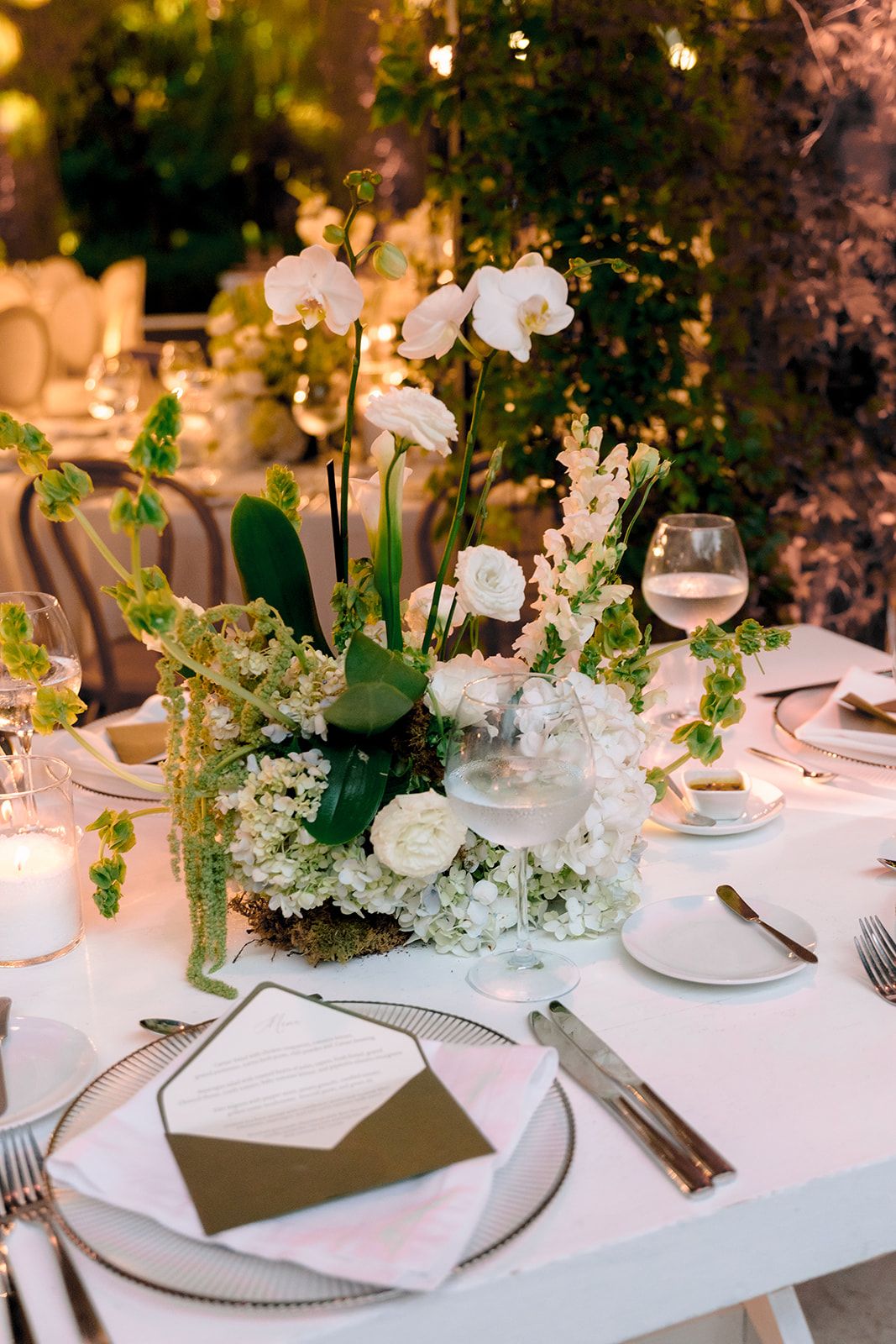 Elegant wedding reception table with white orchid centerpiece, gold accents, crystal glassware, and lush greenery backdrop.
