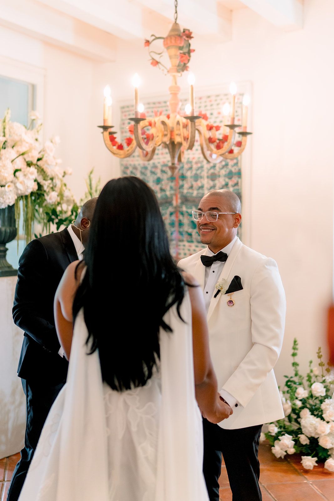 Groom in white tuxedo jacket smiles at bride during indoor wedding ceremony with chandelier and white floral arrangements.