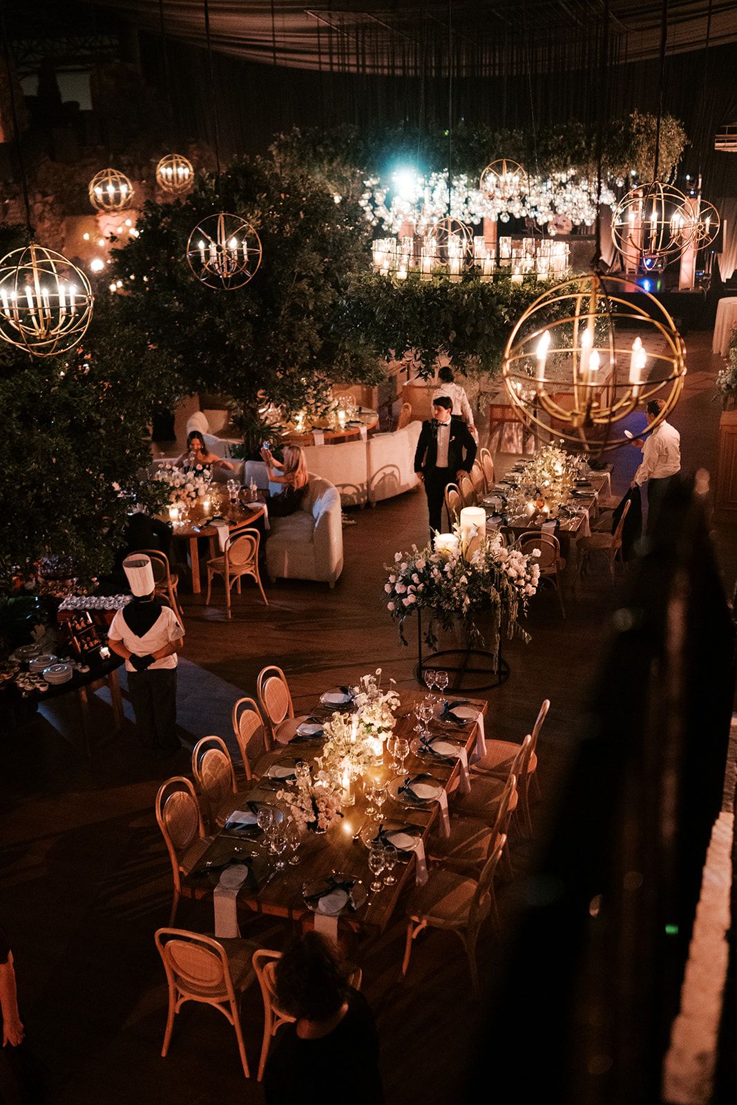 Elegant wedding reception with glowing orb chandeliers, lush greenery, candlelit tables, and wicker chairs in a warmly lit venue.