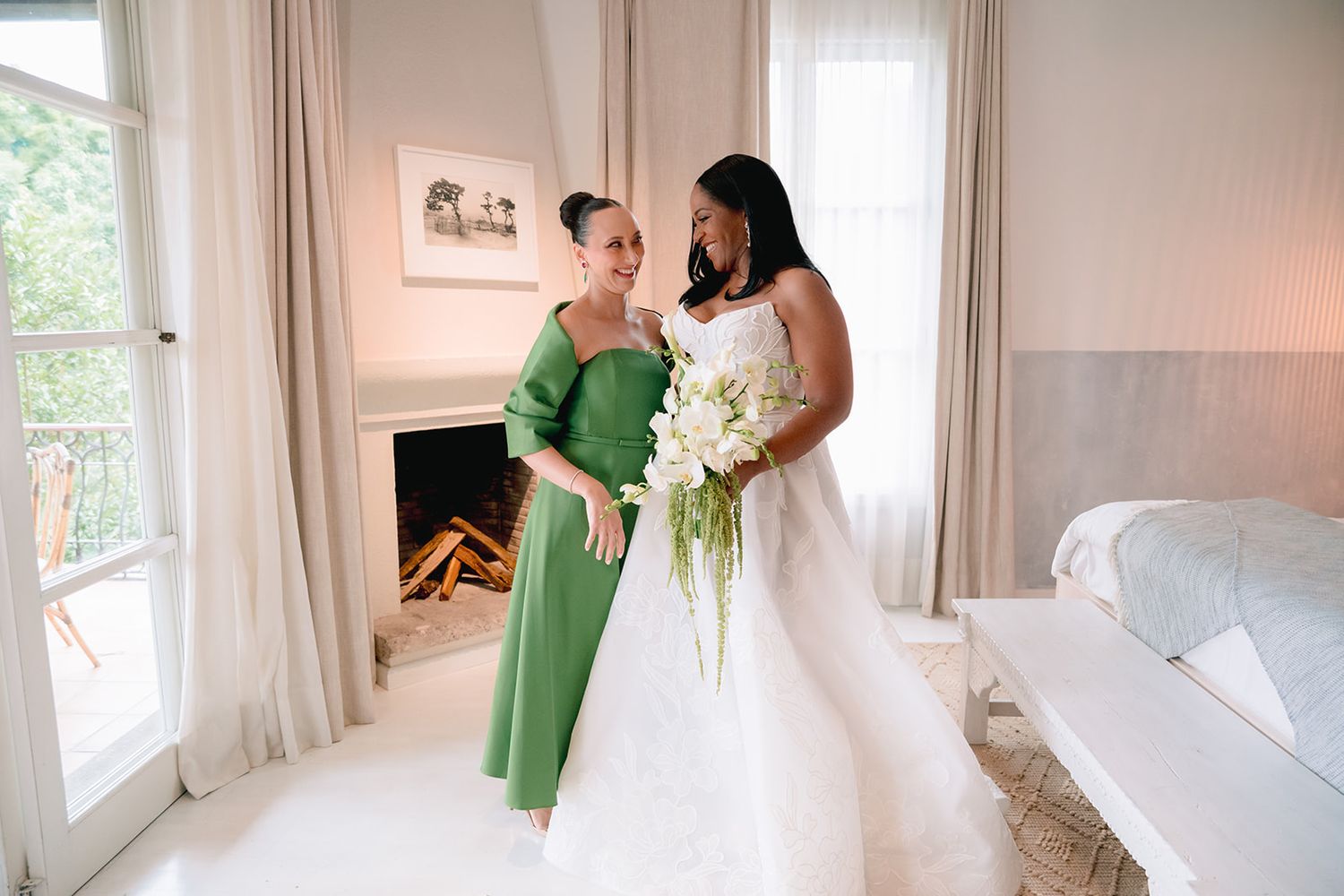 Bride in white gown holds white floral bouquet beside woman in green dress in elegant white room.