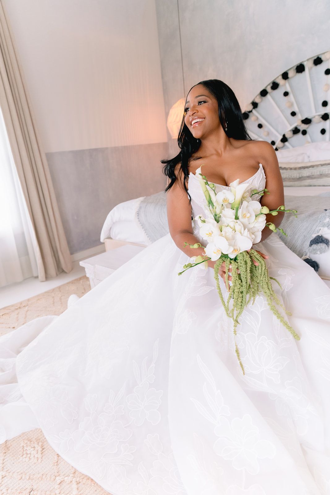 Smiling Black bride in white lace gown holding white floral bouquet, seated on bed in elegant bridal suite.