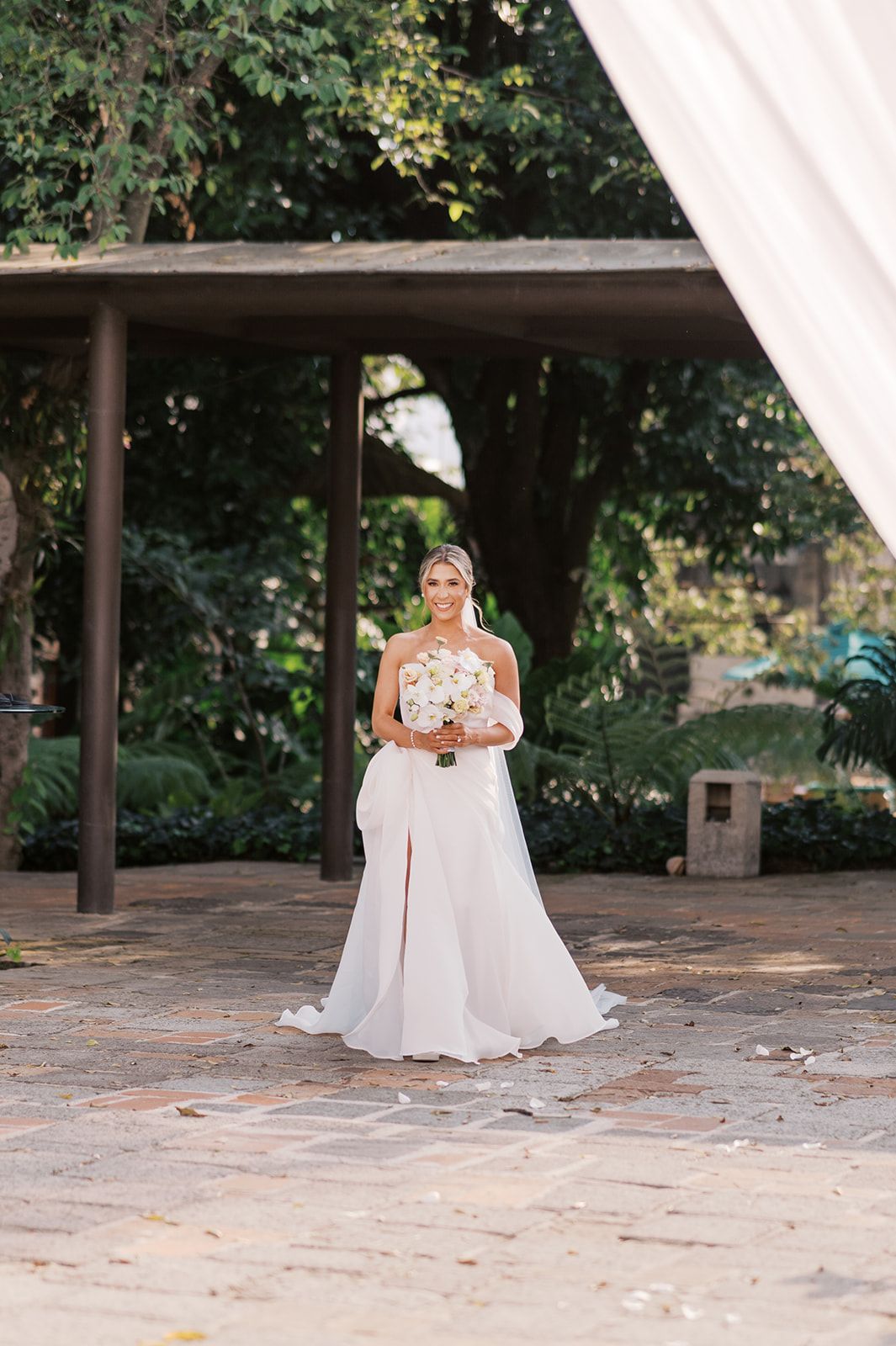 Bride in white halter gown with slit holds bouquet on stone patio surrounded by lush greenery.