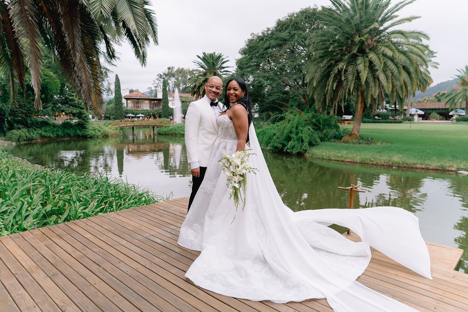 Bride and groom pose on wooden deck by serene pond surrounded by lush tropical greenery on their wedding day.