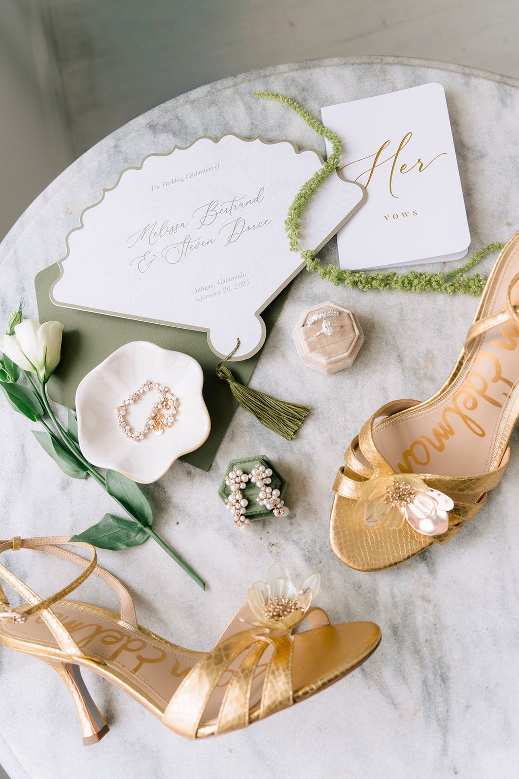 Gold bridal heels, wedding invitation, ring dish, and floral accessories styled on a marble table for an elegant wedding flat lay.
