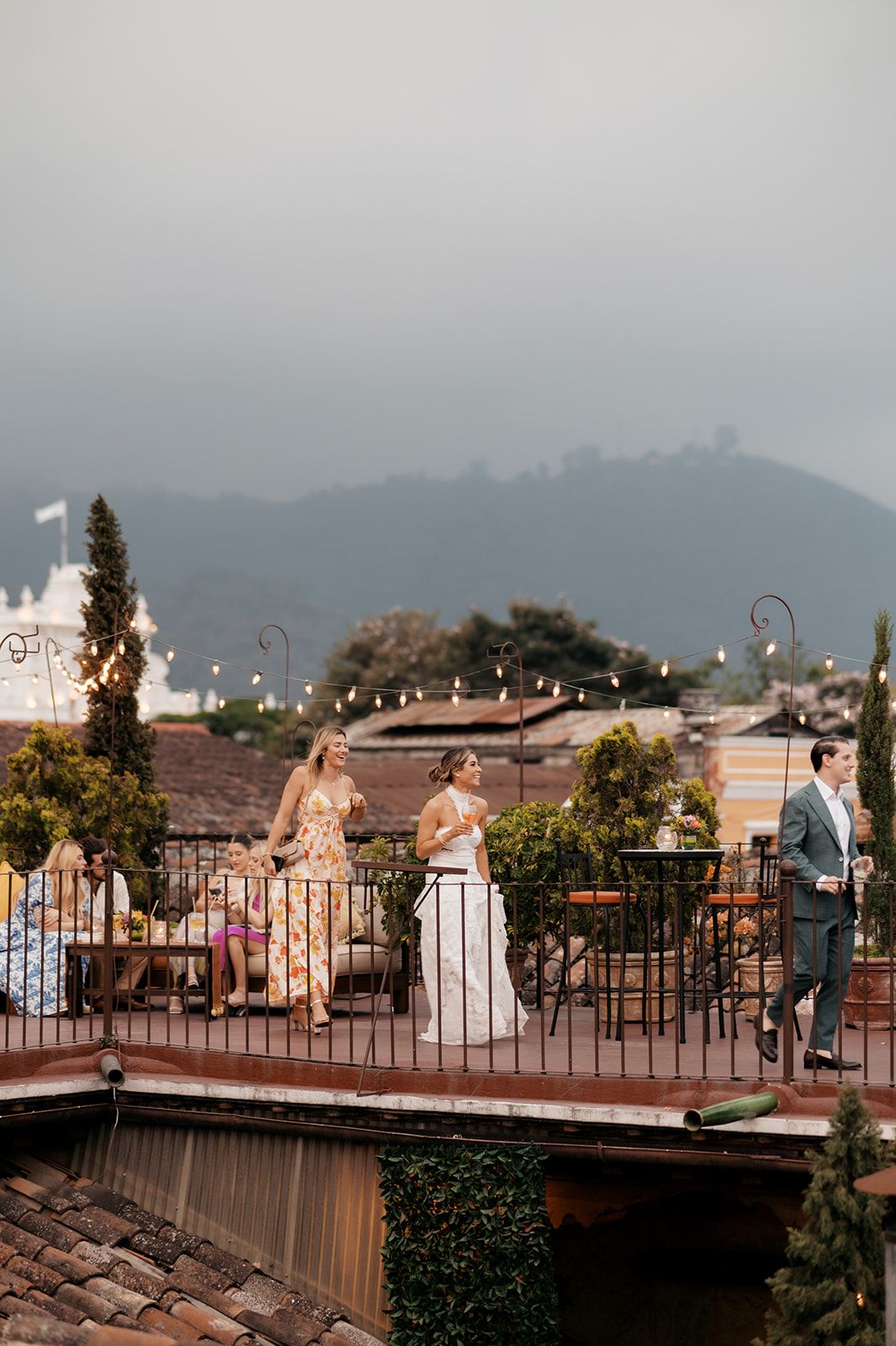 Bride and guests mingle on a rooftop terrace with string lights, overlooking a scenic mountain backdrop.