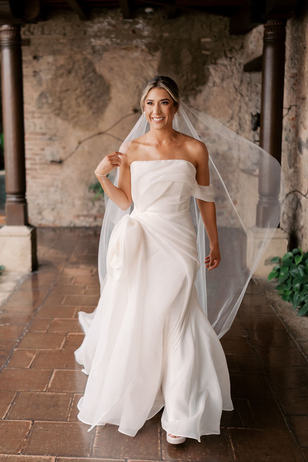 Smiling bride in strapless white gown and flowing veil poses in rustic stone archway.