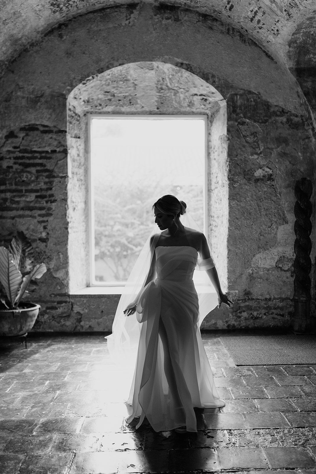 Black and white photo of a bride in a strapless gown standing before an arched window in a rustic stone venue.