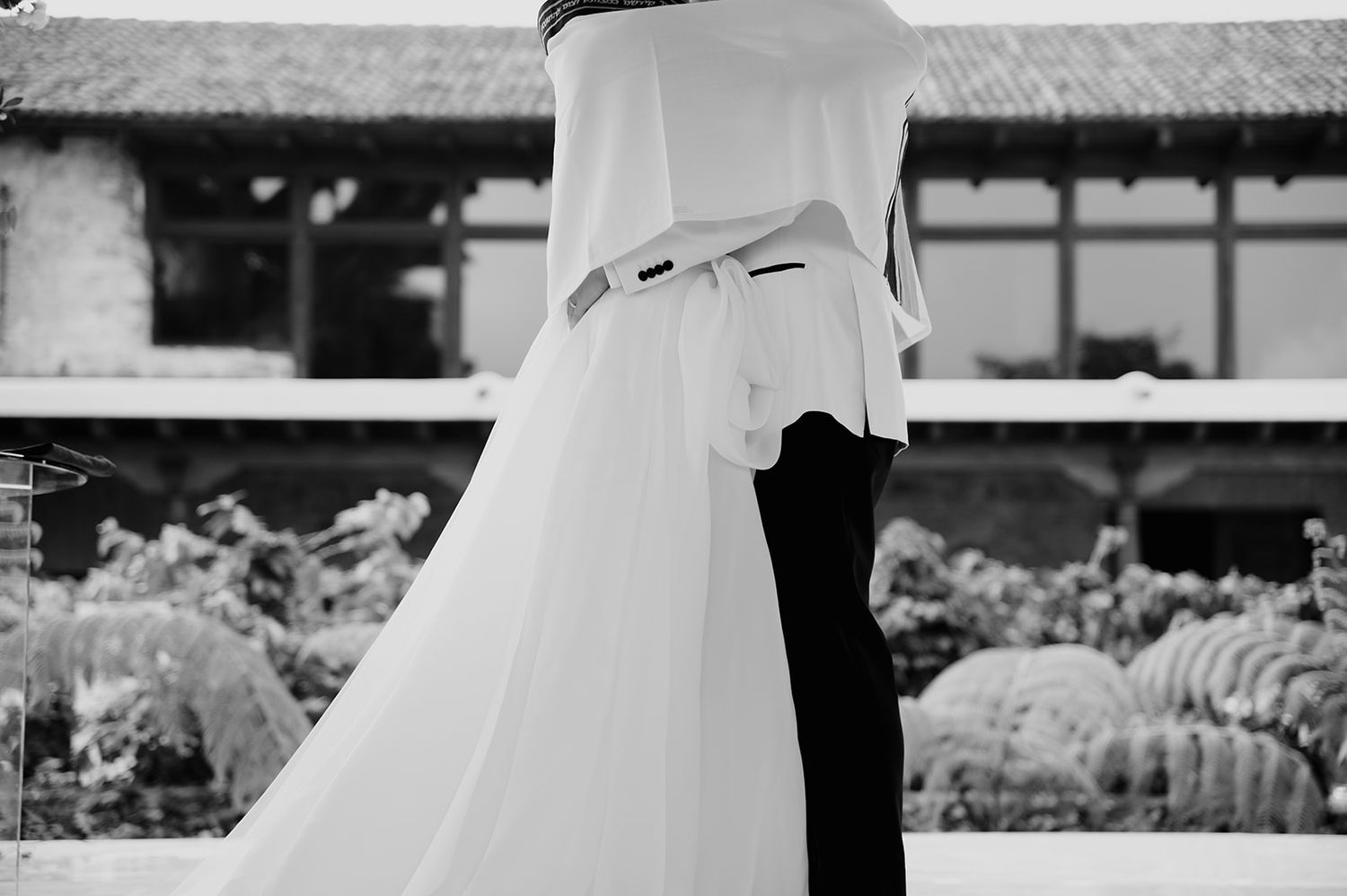 Black and white photo of a couple embracing in wedding attire, with a elegant venue and lush gardens in the background.