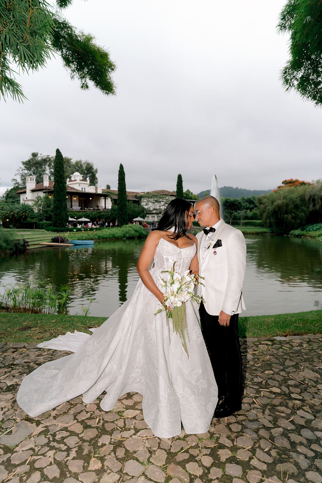 Bride in white gown and groom in white tuxedo share romantic moment by serene lake at elegant outdoor wedding venue.