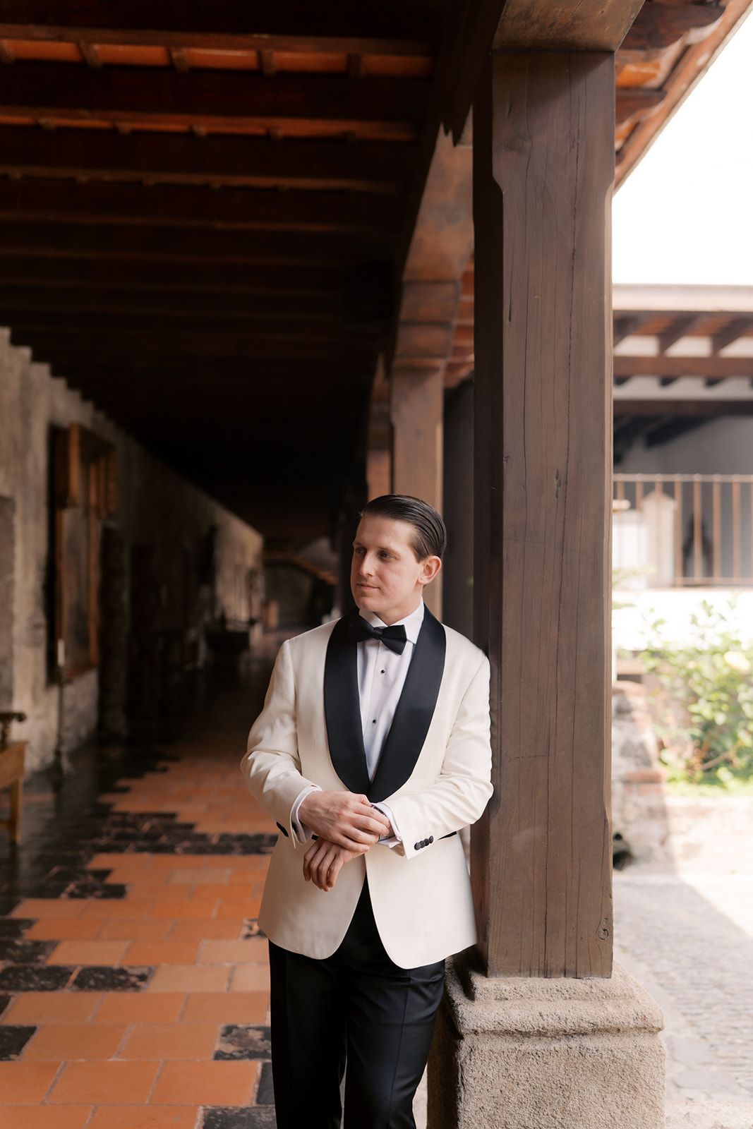 Groom in white tuxedo jacket with black lapels and bow tie poses in rustic colonial corridor.