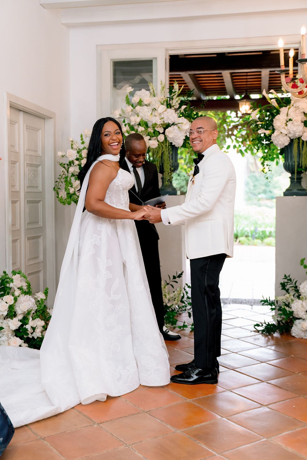 Bride and groom holding hands at elegant wedding ceremony with white floral arrangements and officiant in background.