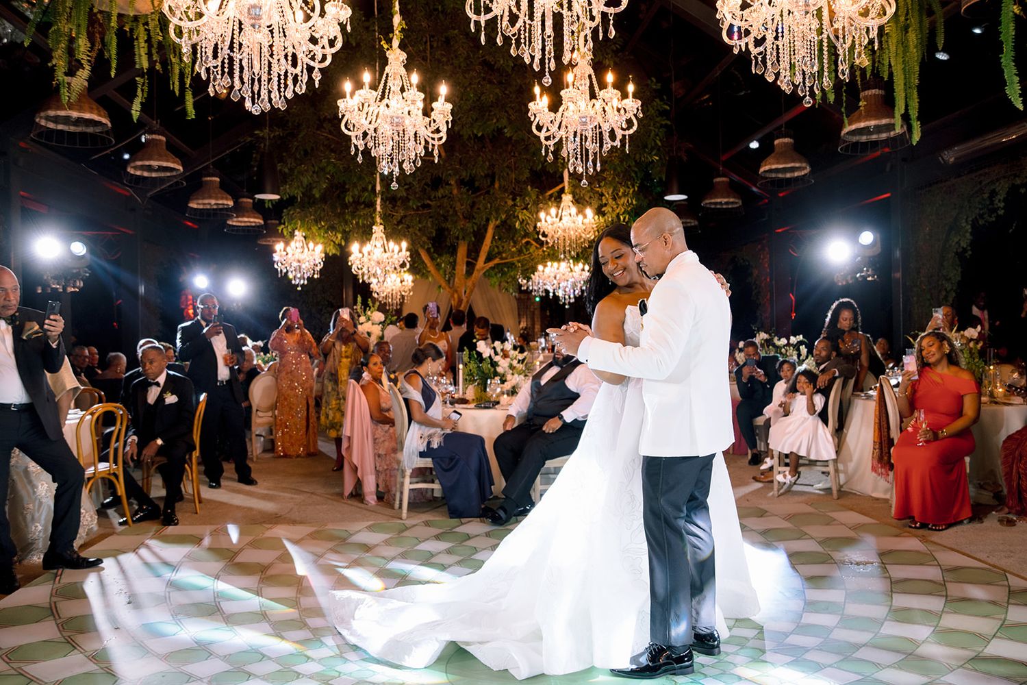 Bride and groom share first dance under glowing chandeliers at elegant outdoor wedding reception with guests watching.