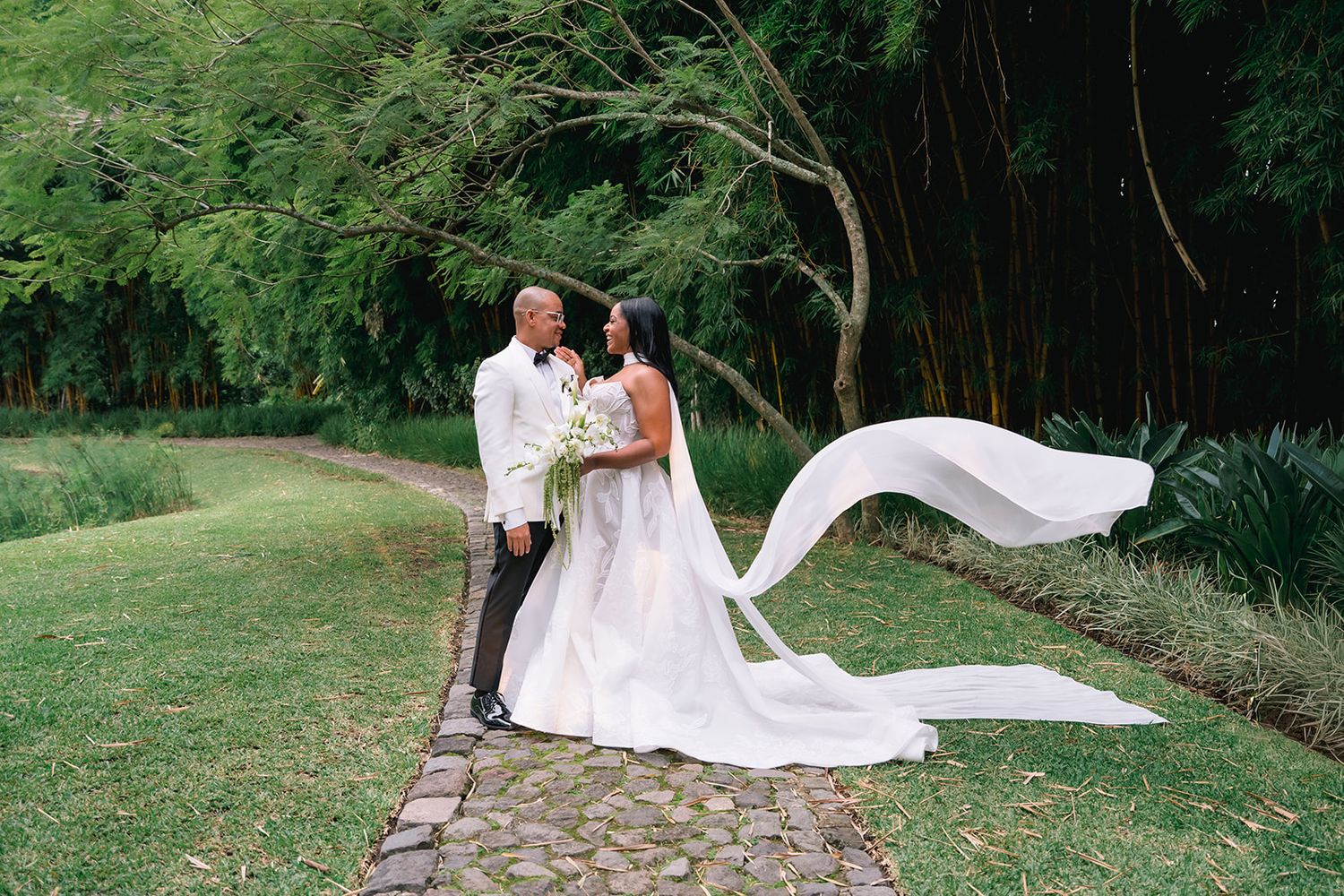 Bride and groom pose romantically on a lush green garden path with a flowing cathedral veil.