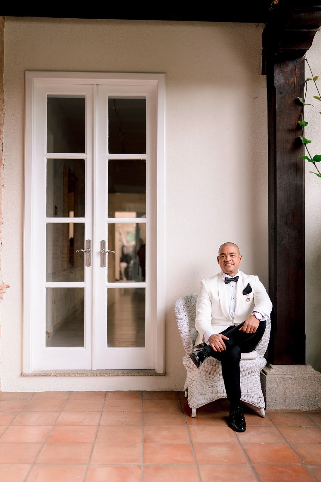 Groom in white tuxedo jacket and bow tie sits in wicker chair beside French doors at elegant venue.