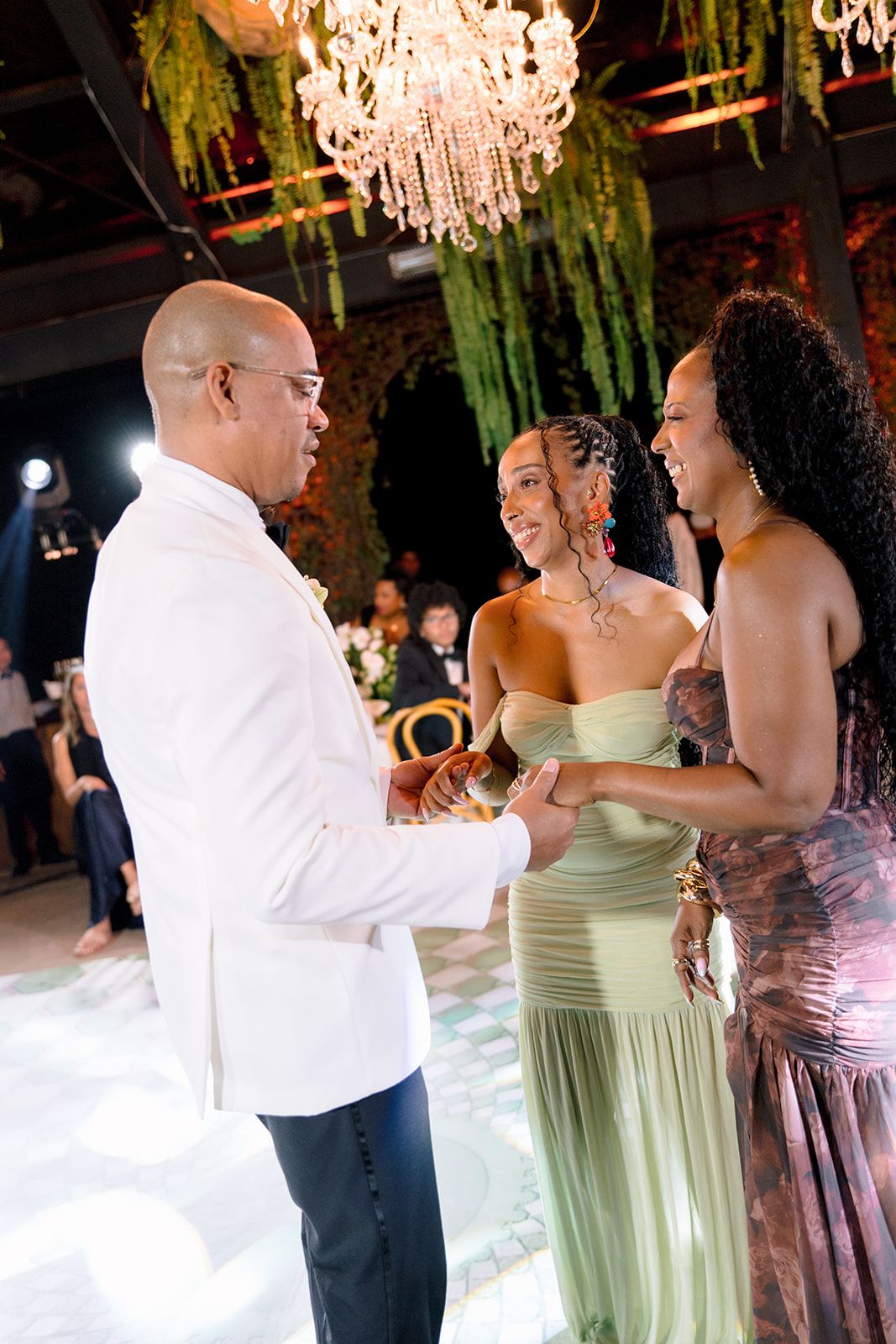 Elegant wedding reception with guests in formal attire dancing under a chandelier with lush greenery decor.